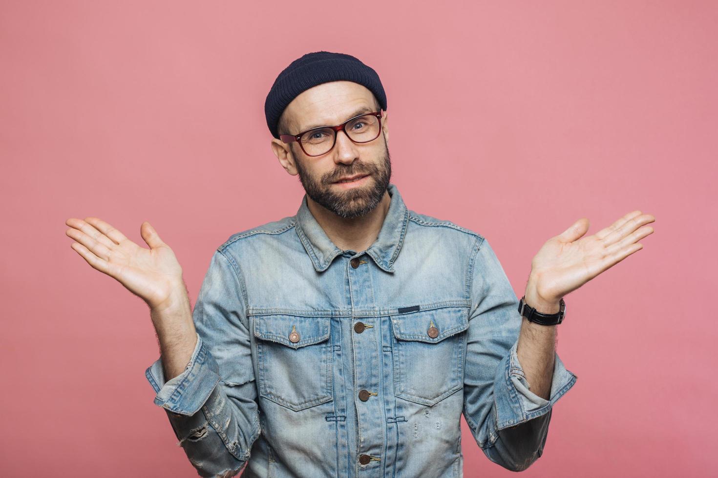 retrato de hombre barbudo vacilante con expresión dudosa, se encoge de hombros, lleva chaqueta vaquera, gafas y sombrero, aislado sobre fondo de estudio rosa. hombre de mediana edad confundido posa en interiores foto