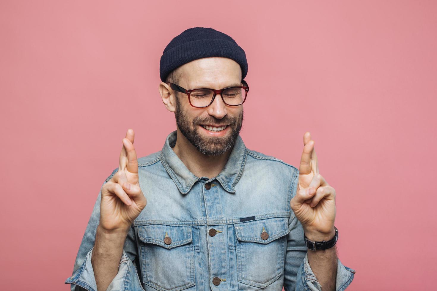 Positive unshaven male with glad look crosses fingers, keeps eyes closed, has great desire and hope for better, wears fashionable clothes, isolated over pink background. People and desire concept photo