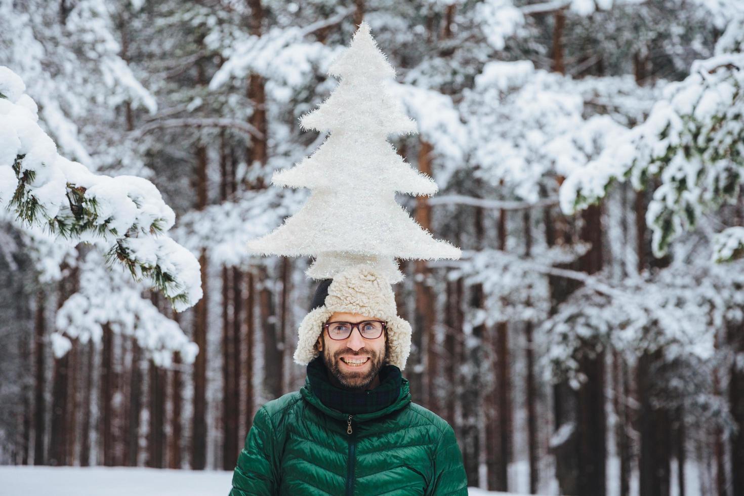 Horizontal shot of smiling male model holds fir tree on head, stands outdor in winter forest, enjoys calmness and tranquil atmosphere. Caucasian unshaven man with cheerful expression photo