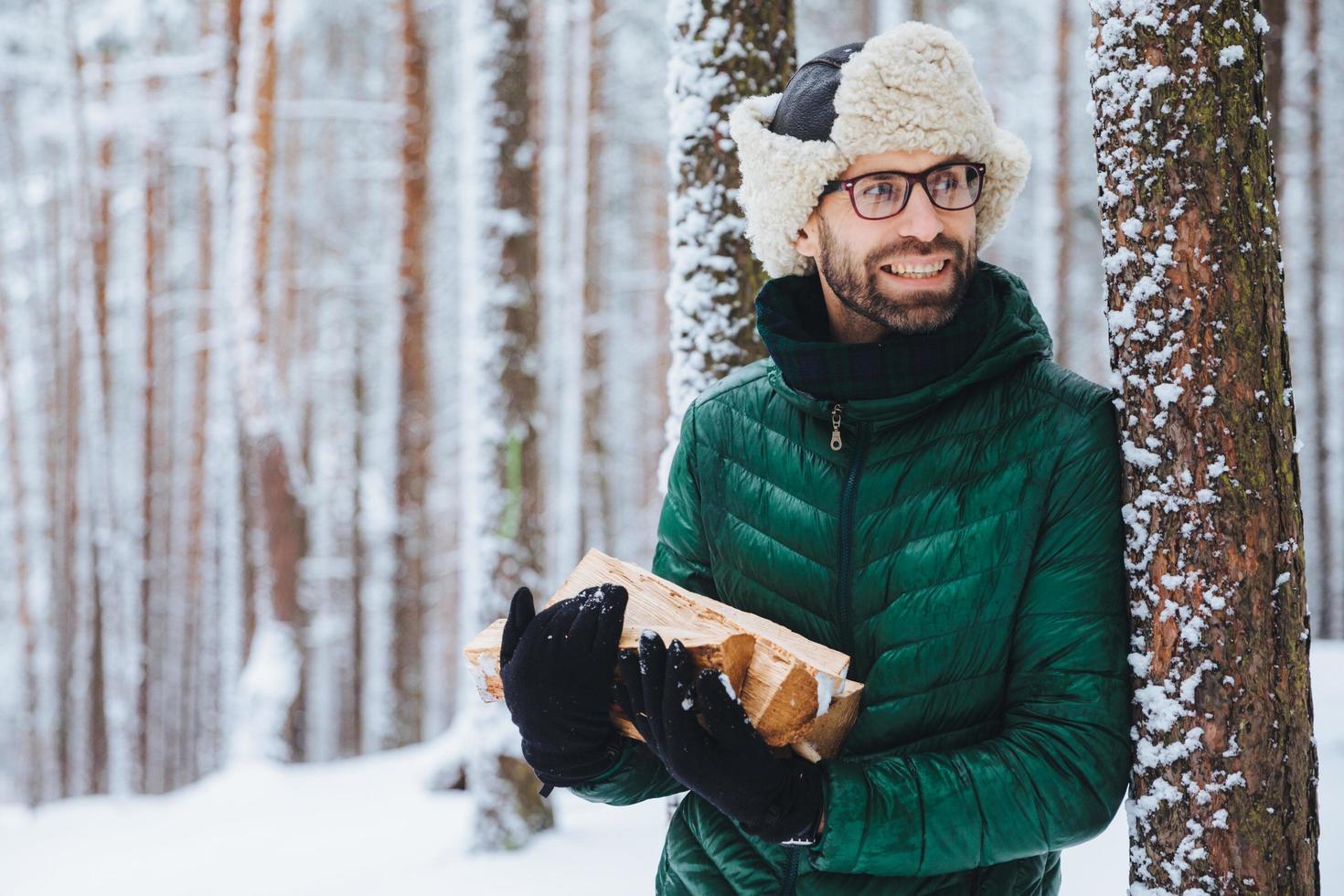 el joven barbudo usa un sombrero cálido y una chaqueta verde, sostiene un montón de leña, mira felizmente a un lado, nota algo, le gusta el invierno y el clima helado. hombre atractivo soñador pasa tiempo en el bosque foto