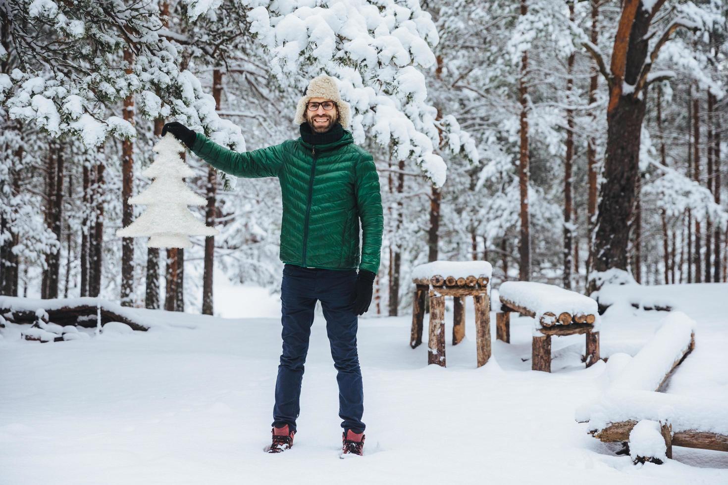 Full growth portrait of cheerful smiling male demonstrates white artificial fir tree, stands in winter forest has happy expression, glad to spend time outdoors. Winter and season concept photo