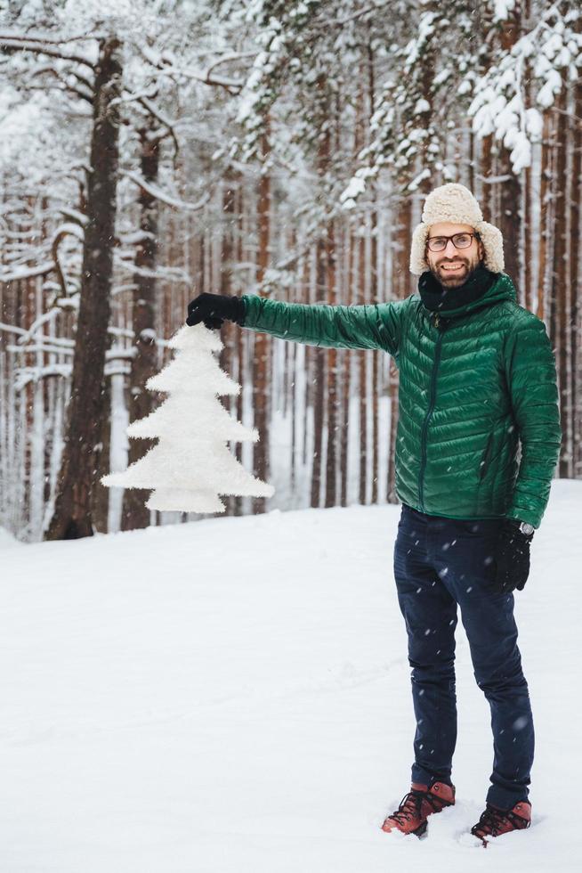 Vertical portrait of positive smiling male wears fashionable warm clothes, keeps white fir tree in hands, poses in white winter forest. Happy unshaven attractive man spend free time or winter holidays photo