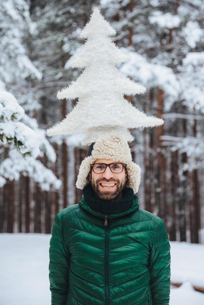 Vertical portrait of cheerful bearded man has fun alone in winter forest, keeps artificial fir tree, poses outdoors, admires frosty snowy weather, expresses positiveness and pleasant emotions photo