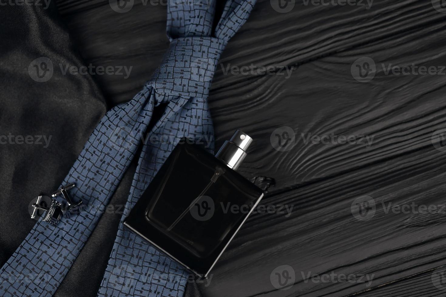 A bottle of mens cologne and cufflinks with blue tie lie on a black luxury fabric background on a wooden table. Mens accessories photo