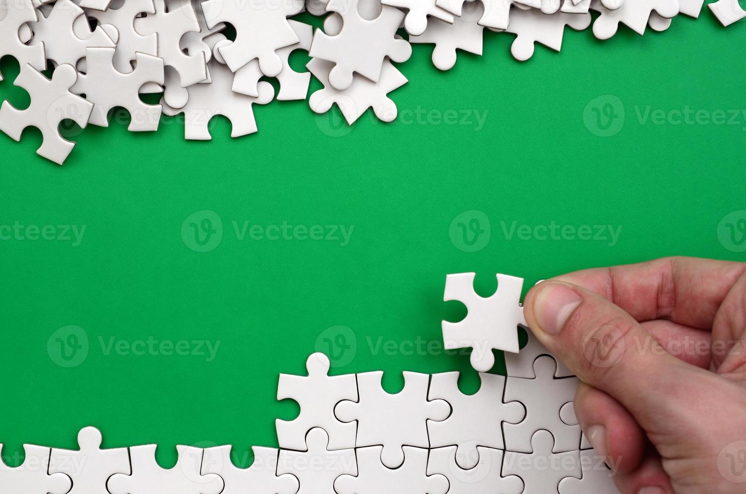 The hand folds a white jigsaw puzzle and a pile of uncombed puzzle pieces lies against the background of the green surface. Texture photo with space for text