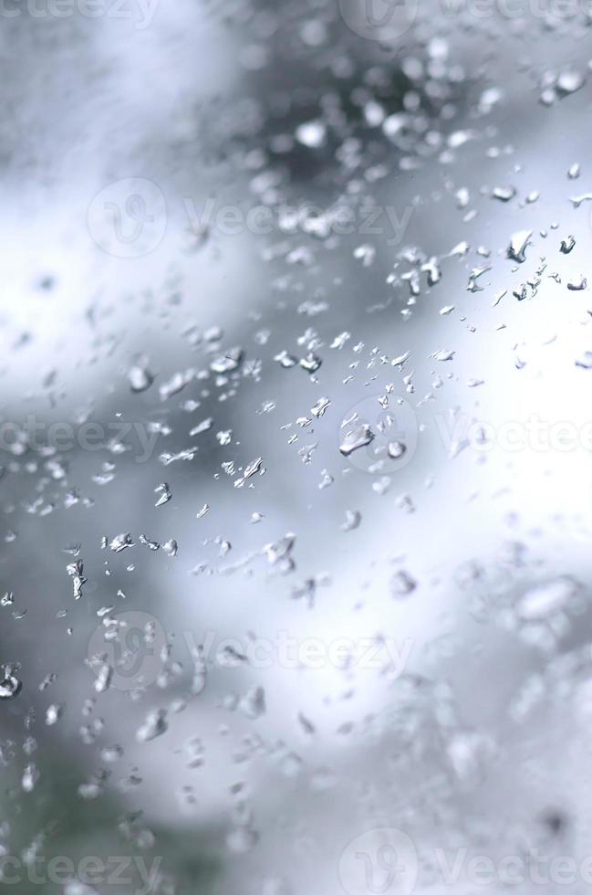 A photo of rain drops on the window glass with a blurred view of the blossoming green trees. Abstract image showing cloudy and rainy weather conditions