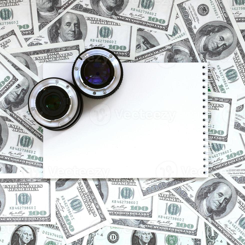 Two photographic lenses and white notebook lie on the background of a lot of dollar bills. Space for text photo