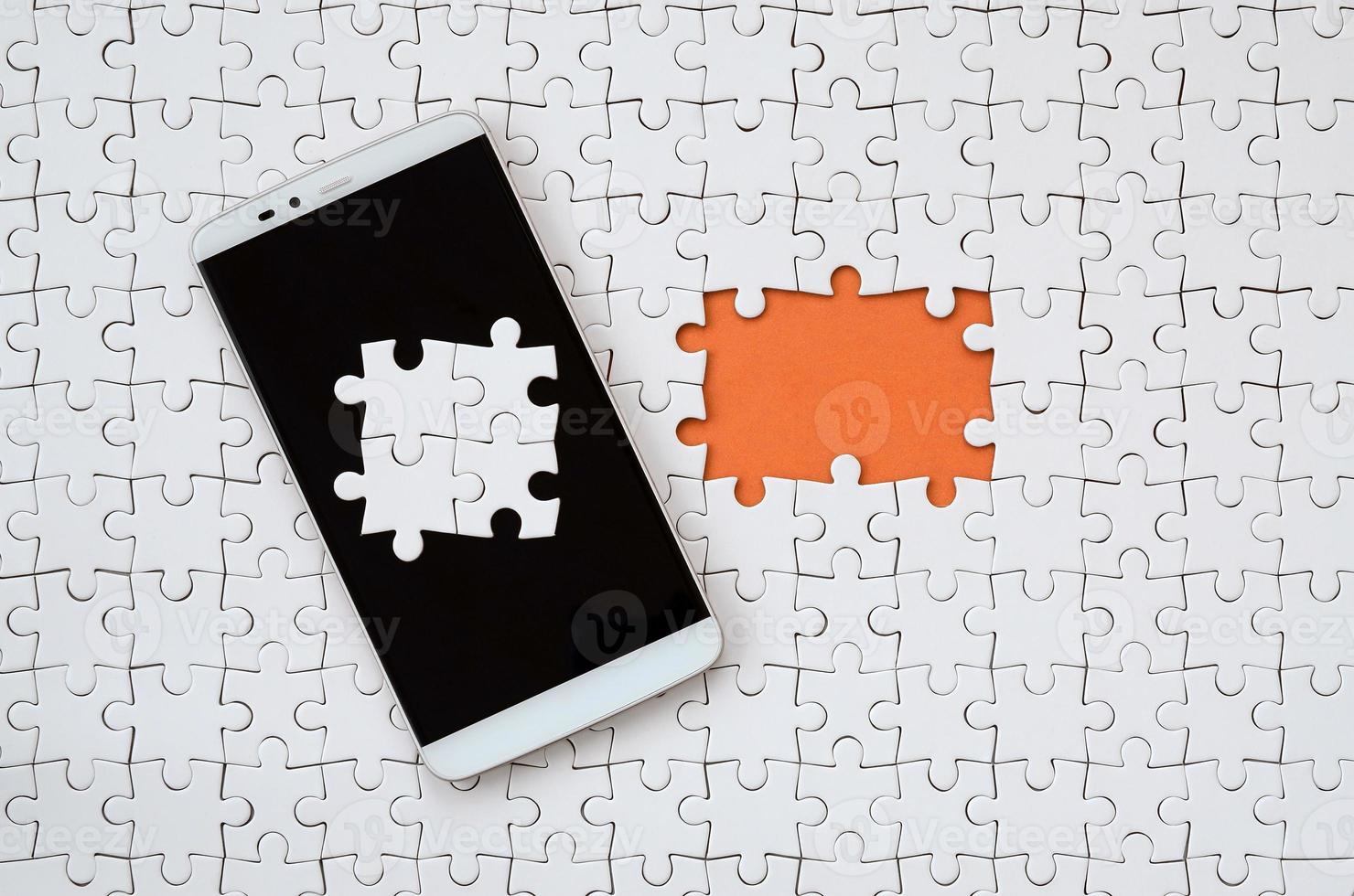 A modern big smartphone with several puzzle elements on the touch screen lies on a white jigsaw puzzle in an assembled state with missing elements photo