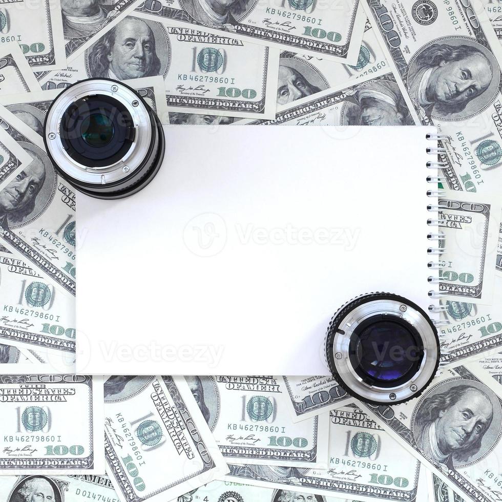 Two photographic lenses and white notebook lie on the background of a lot of dollar bills. Space for text photo