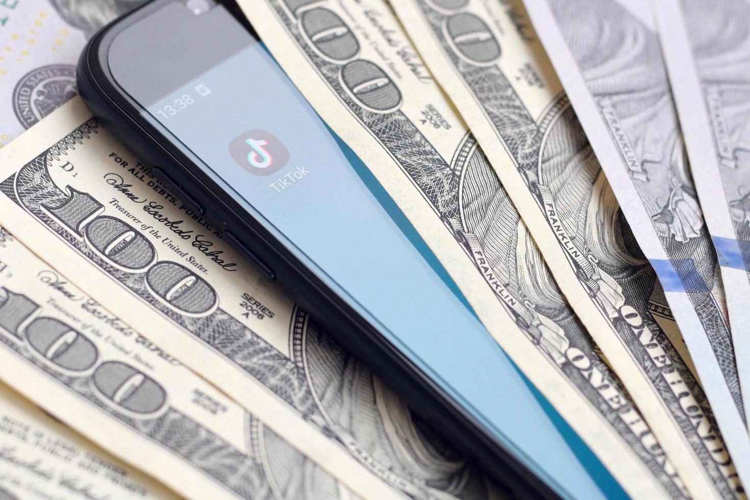 KHARKOV, UKRAINE - JULY 2, 2021 Smartphone screen with Tiktok app and lot of hundred dollar bills. Business and social networking concept photo