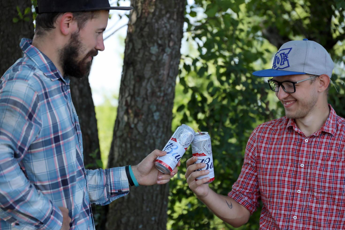 SUMY, UKRAINE - AUGUST 01, 2021 Young man raise Budweiser Bud beer can with male friend on blurred river with kayak and trees. Budweiser is one of the most popular beer brands in the USA photo