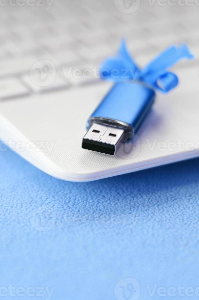 Brilliant blue usb flash memory card with a blue bow lies on a blanket of soft and furry light blue fleece fabric beside to a white laptop. Classic female gift design for a memory card photo