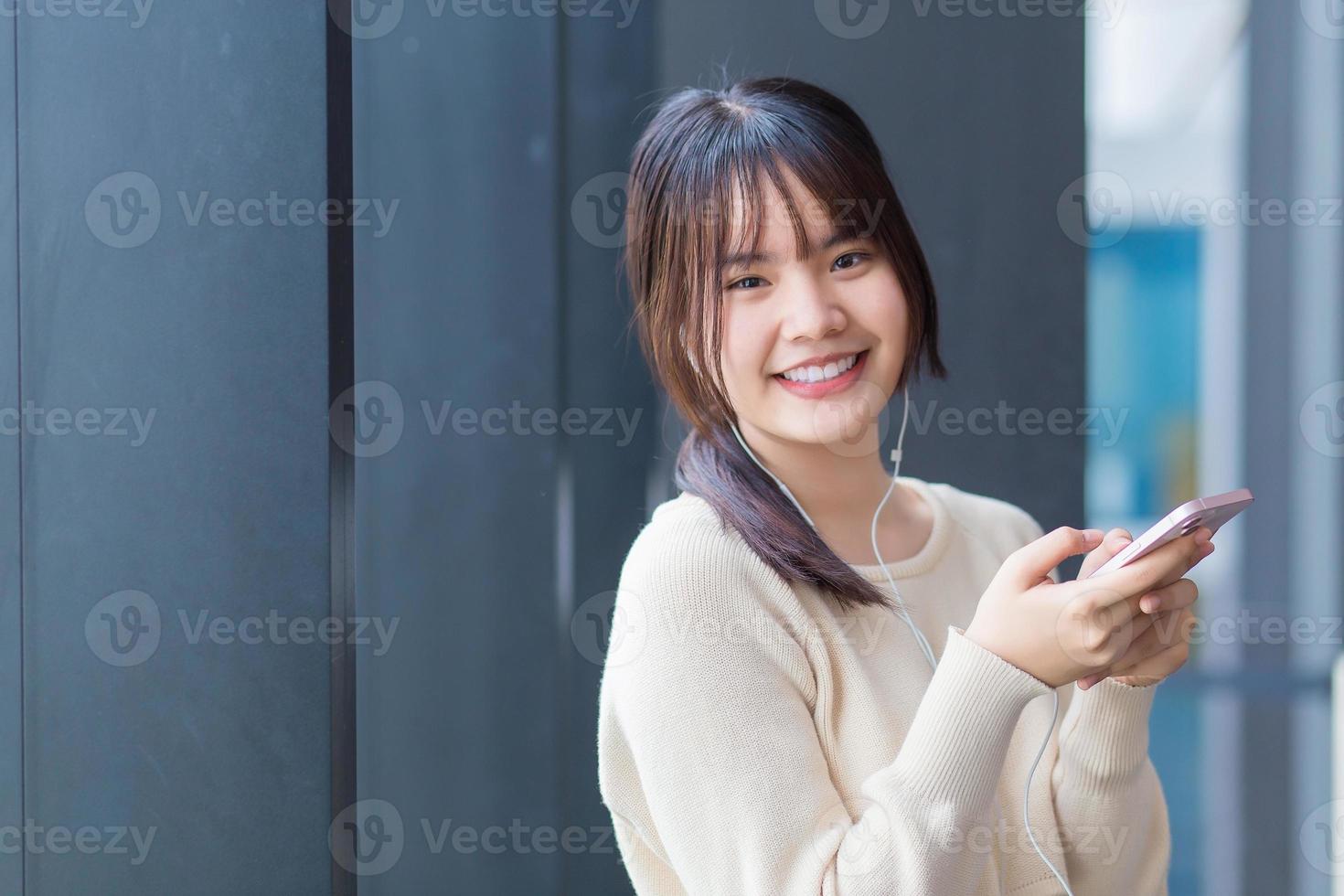 Cute teenage Asian student wearing long-sleeved shirt standing confidently is using her smartphone and headphones to make fun video call with her friends as she waits to enter the classroom at school. photo