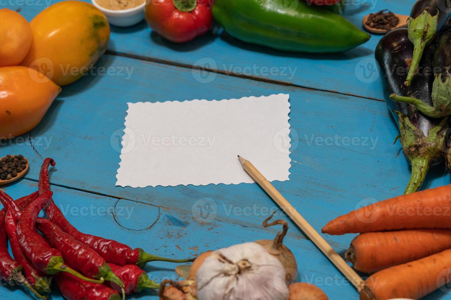 las verduras se disponen alrededor de una hoja de papel y un lápiz. espacio vacío para texto. verduras, en blanco vacío para la receta sobre un fondo azul. foto