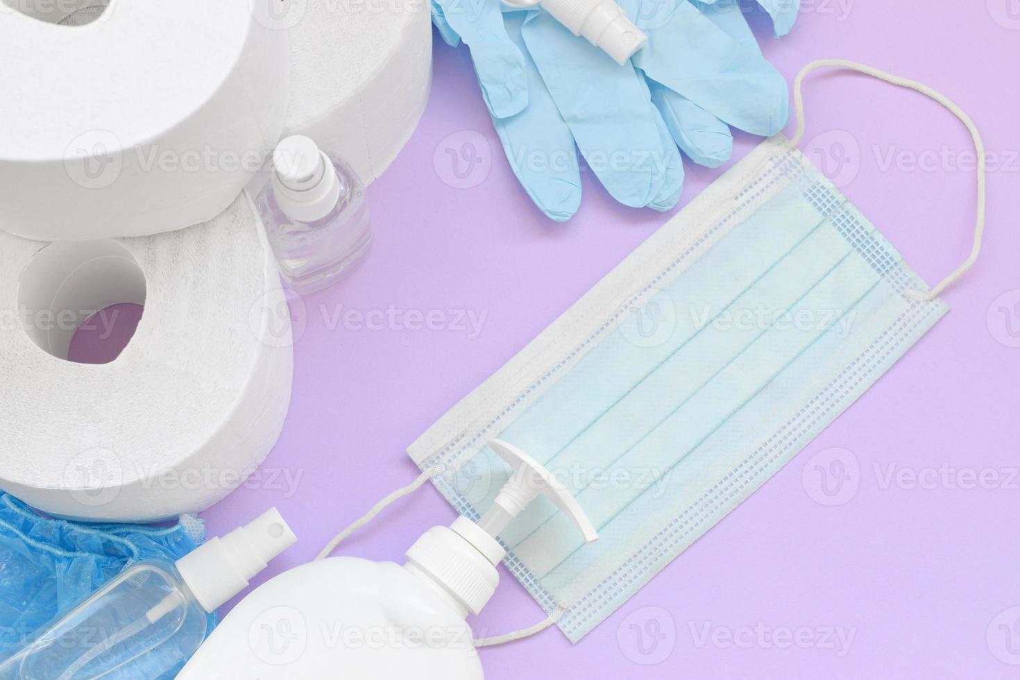 Set of important items for Covid-19 quarantine. Toilet paper, rubber disposable gloves with surgical face mask and hand sanitizer with liquid soap bottle on lilac background photo