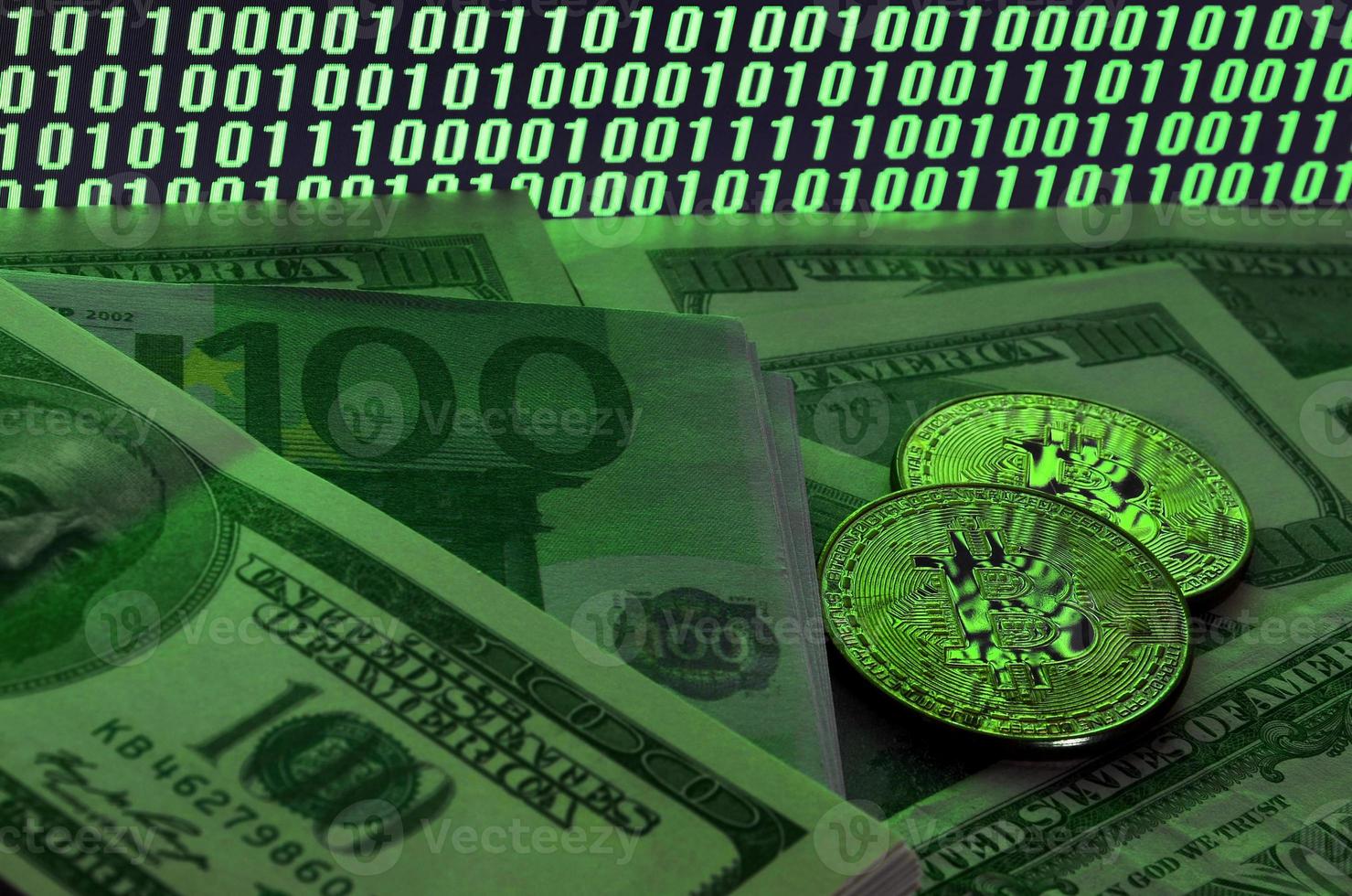 Two bitcoins lies on a pile of dollar bills on the background of a monitor depicting a binary code of bright green zeros and one units on a black background. Low key lighting photo