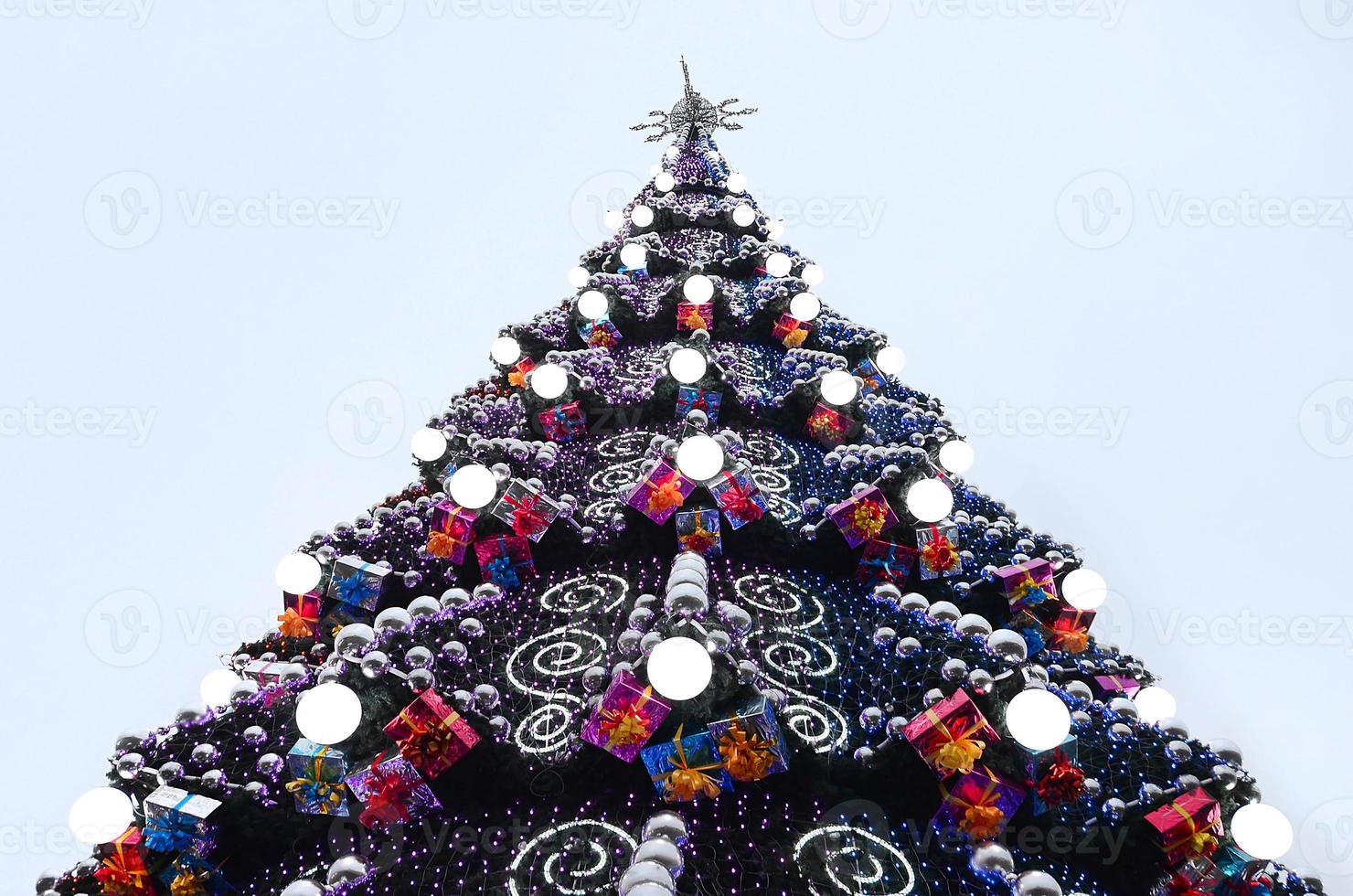 A fragment of a huge Christmas tree with many ornaments, gift boxes and luminous lamps. Photo of a decorated Christmas tree close-up with copy space