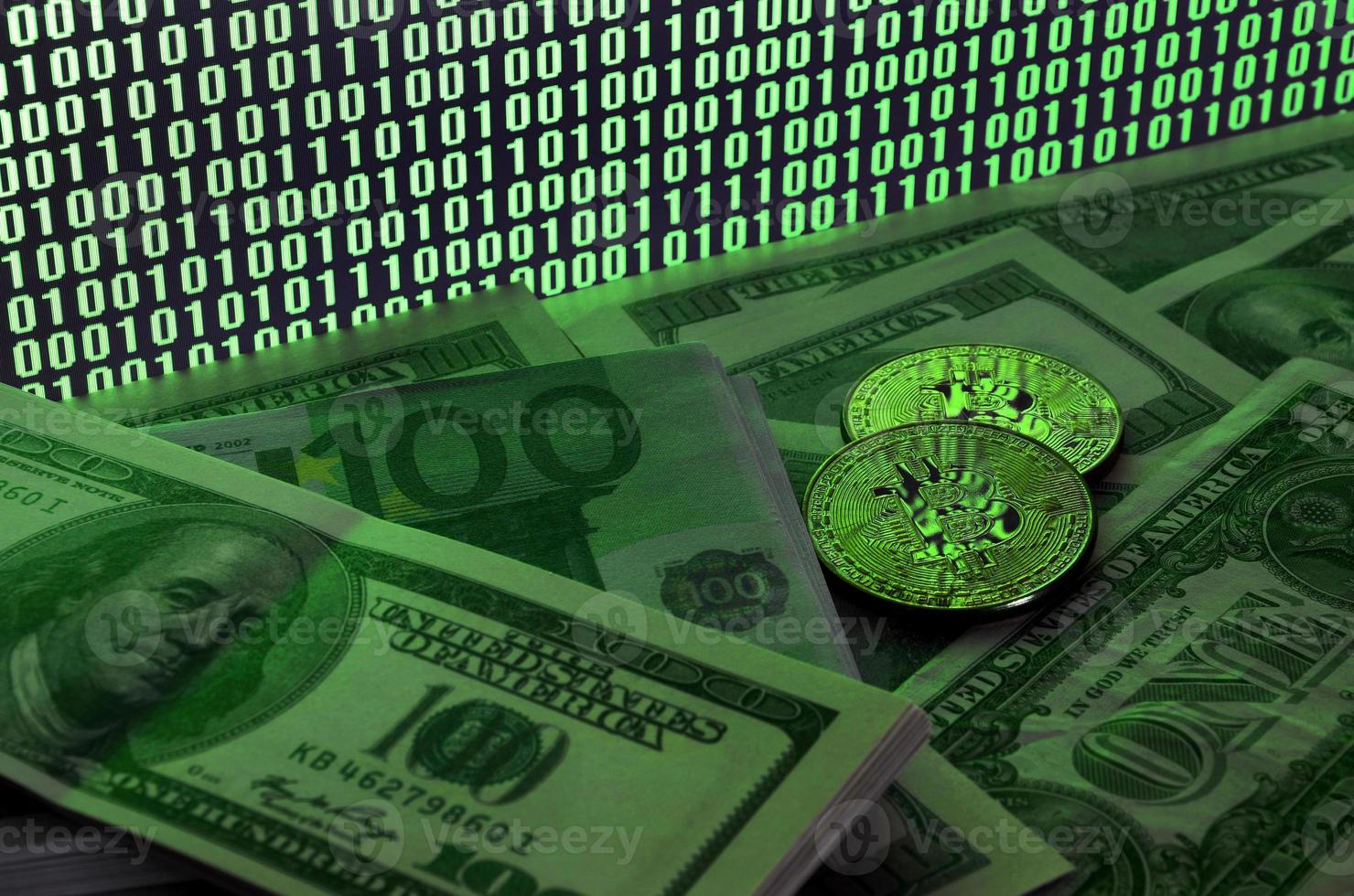 Two bitcoins lies on a pile of dollar bills on the background of a monitor depicting a binary code of bright green zeros and one units on a black background. Low key lighting photo