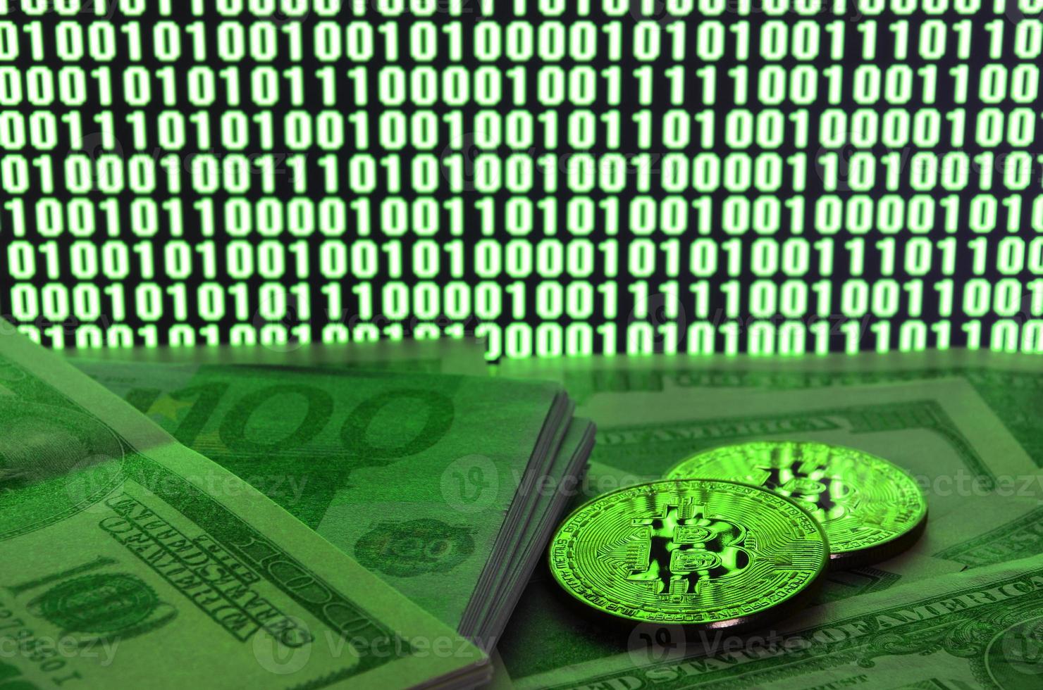 Two bitcoins lies on a pile of dollar bills on the background of a monitor depicting a binary code of bright green zeros and one units on a black background. Low key lighting photo