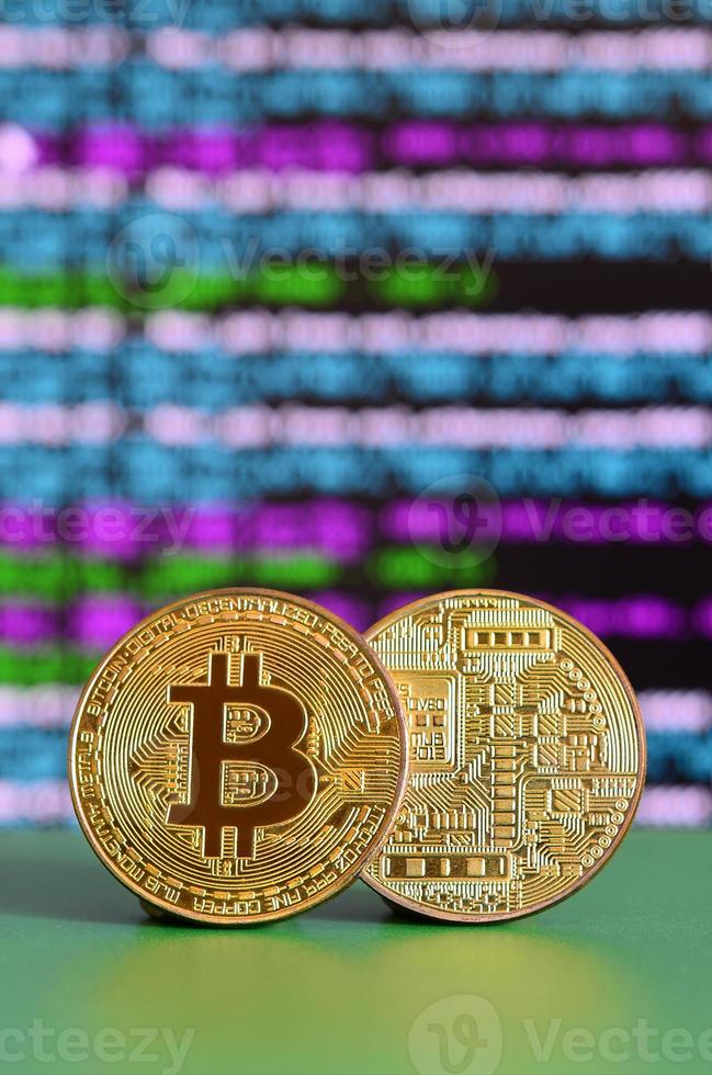 Two gold bitcoins lie on the green surface on the background of the display, which shows the process of mining the crypto currency photo