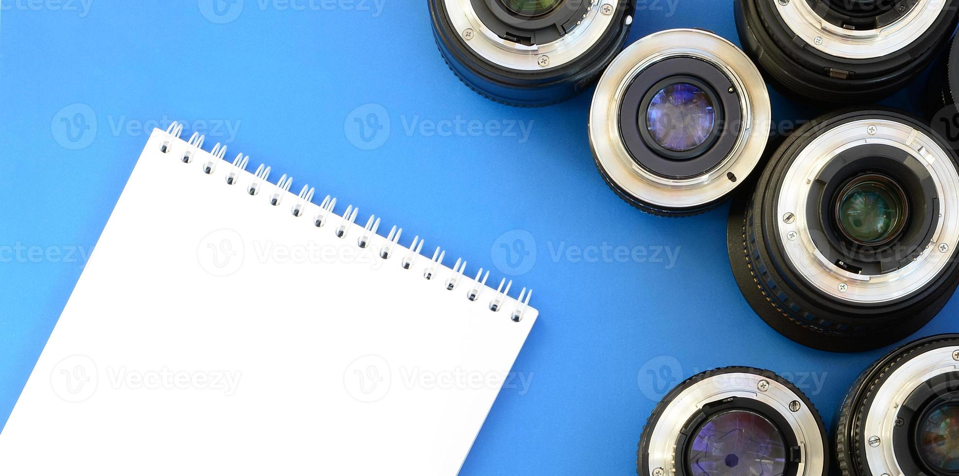 varias lentes fotográficas y un cuaderno blanco yacen sobre un fondo azul brillante. espacio para texto foto