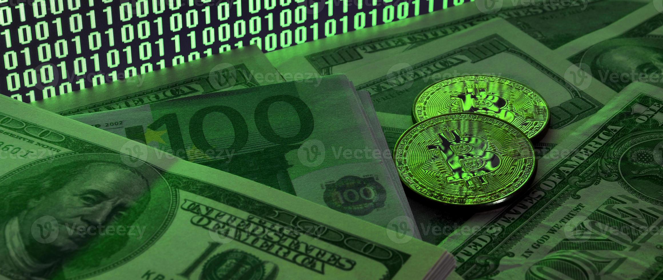 Two bitcoins lies on a pile of dollar bills on the background of a monitor depicting a binary code of bright green zeros and one units on a black background. Low key lighting photo