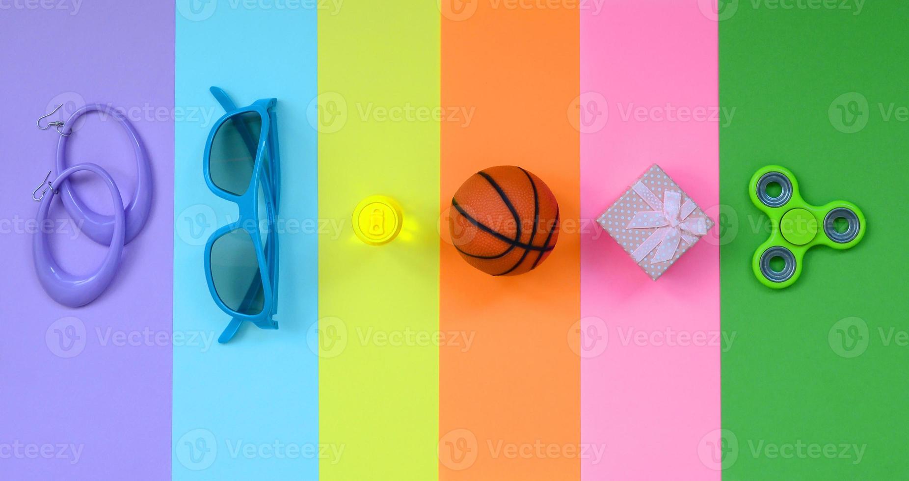 Trendy fashionable pastel composition with earrings, sunglasses, beverage can, basketball ball, gift box and spinner on background of pink, violet, green, orange, yellow and blue colors photo