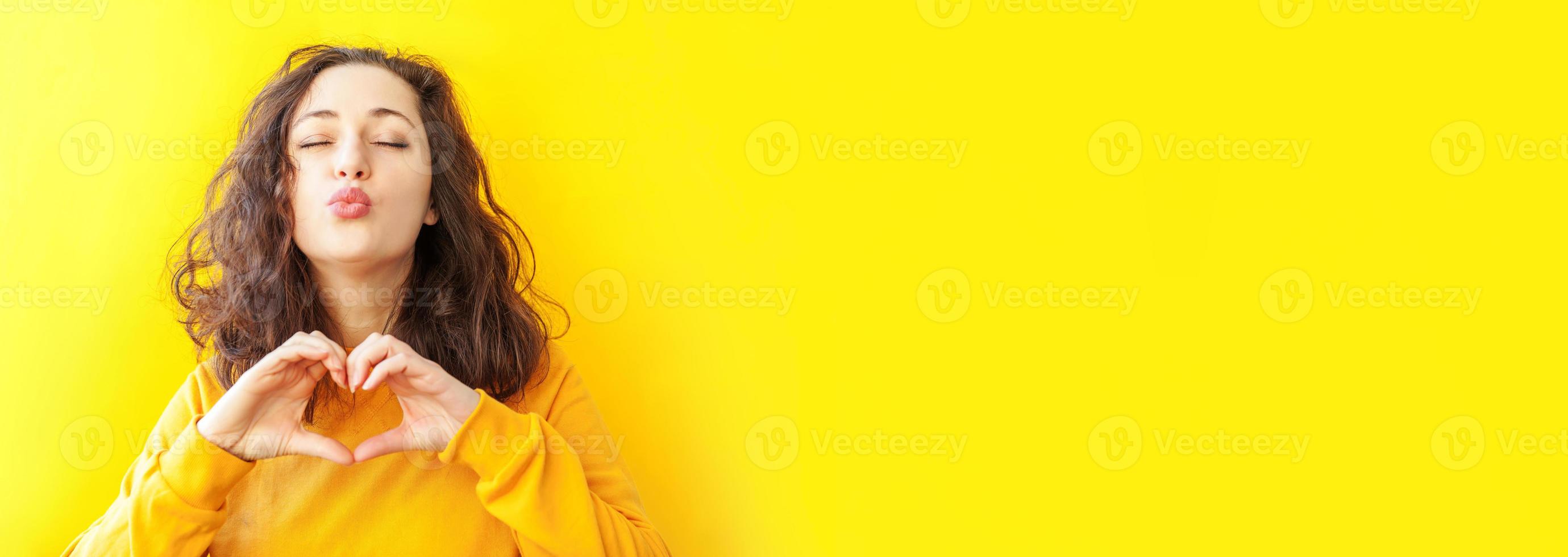 Love heart shape peace. Beauty portrait young happy woman showing heart sign with hands on yellow background isolated. European girl. Positive human emotion facial expression body language Banner photo
