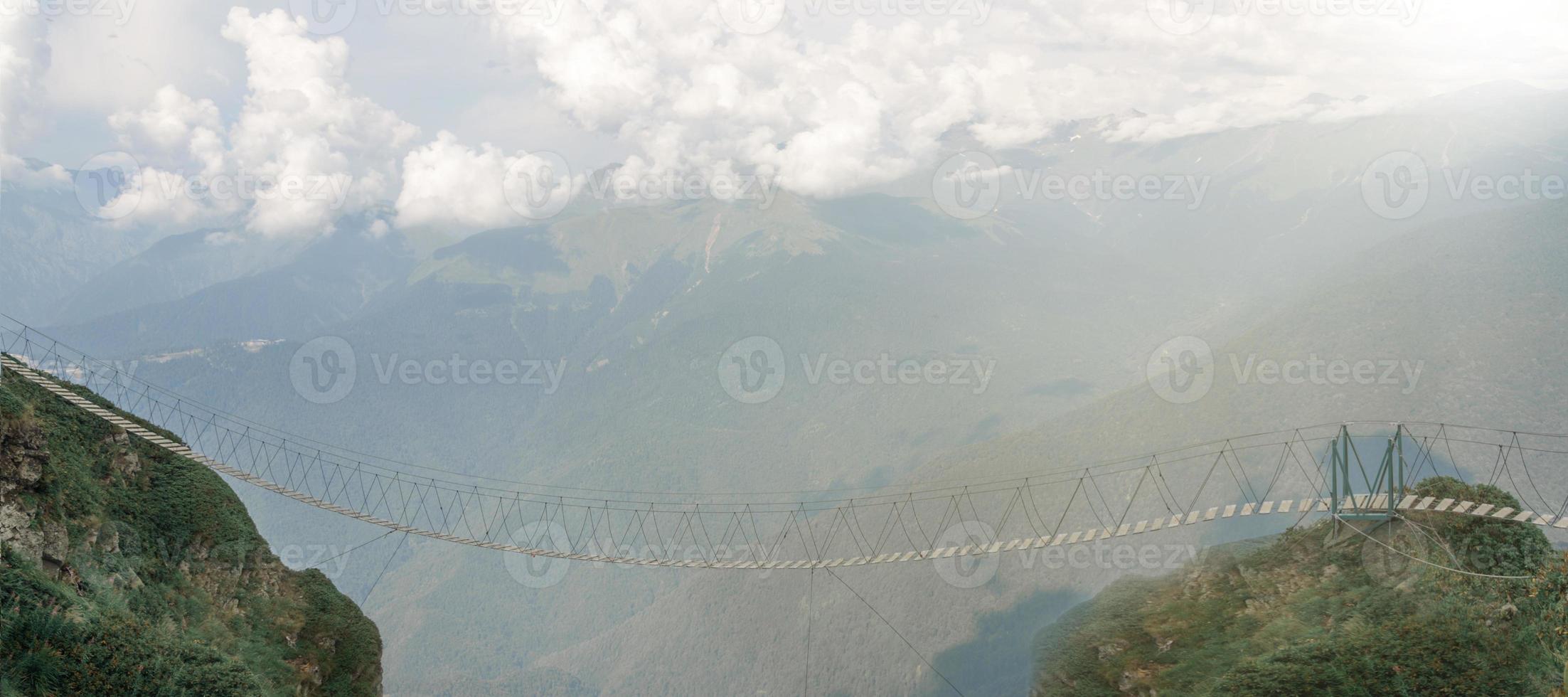 The only way in the mountains lit by sunlight. Empty suspension bridge high in the mountains on the background of clouds. photo