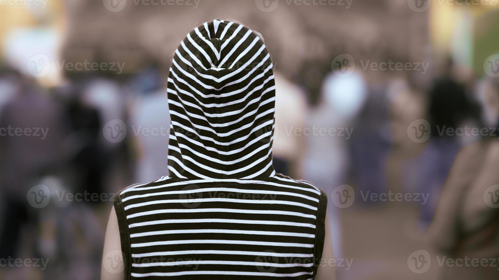 Rear view lonely girl in a vest with a hood stands against the crowd. Background in blur. photo