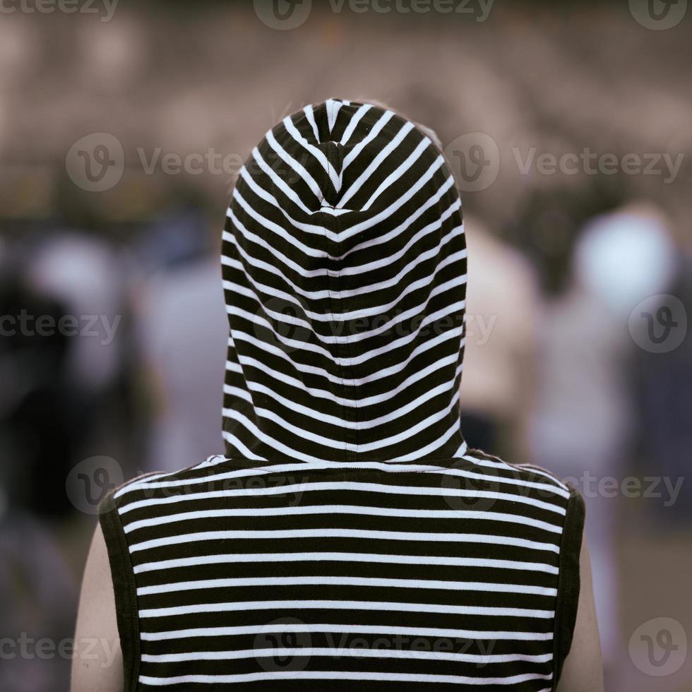 Rear view lonely girl in a vest with a hood stands at a street concert. Background in blur. photo