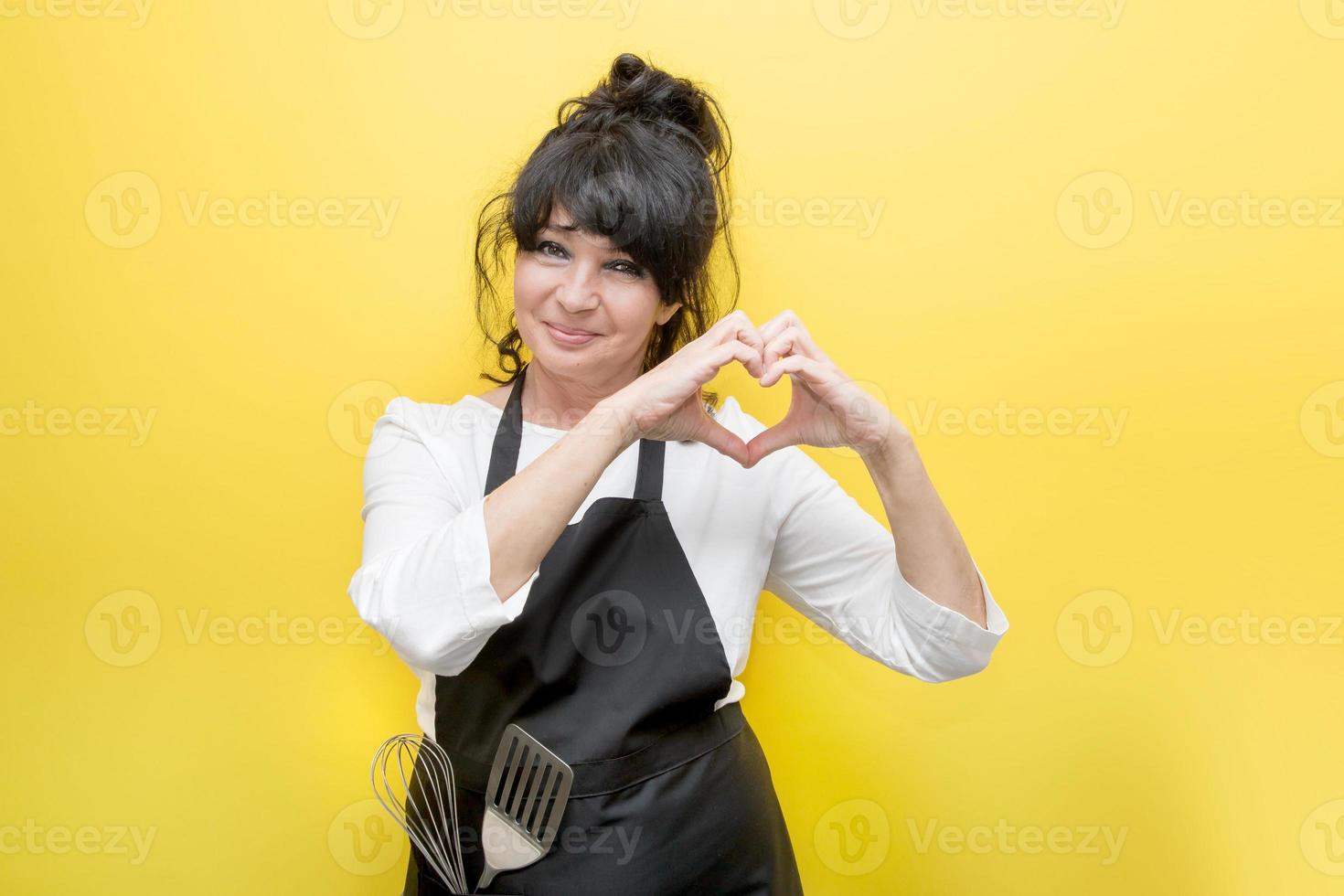 pretty elderly woman in a chef's apron, in whose pocket a whisk and a spatula shows a heart with her hands, on a yellow background, the concept of life is beautiful at any age, love for cooking photo