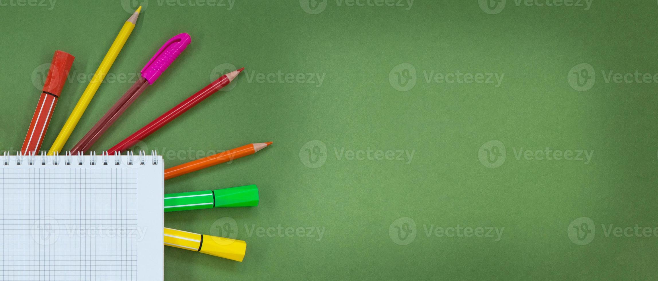 Multicolored pens, pencils, felt tip pens with notepad with cell on side on green background. Concept back to school, learning, drawing. Copy space photo