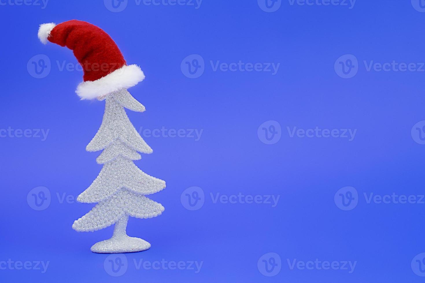 White shiny Christmas tree with Santa Claus hat on its head on blue background. New Year. Copy space photo