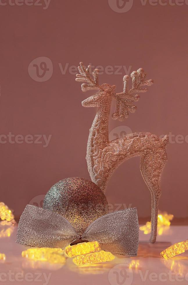 Shiny deer stands next to ball with glitter and bow and Christmas lights on pink background. New Year. Vertical. Copy space photo