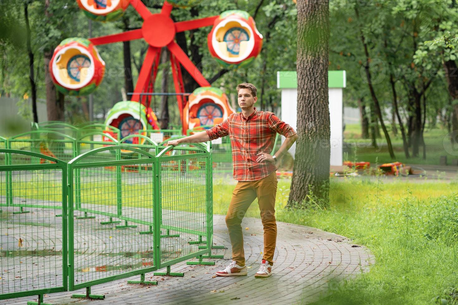 a young man in a plaid shirt is standing in a park with a phone in his hands, behind him is an attraction photo