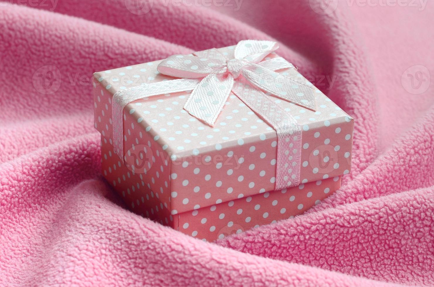 A small gift box in pink with a small bow lies on a blanket of soft and furry light pink fleece fabric with a lot of relief folds. Packing for a gift to your lovely girlfriend photo