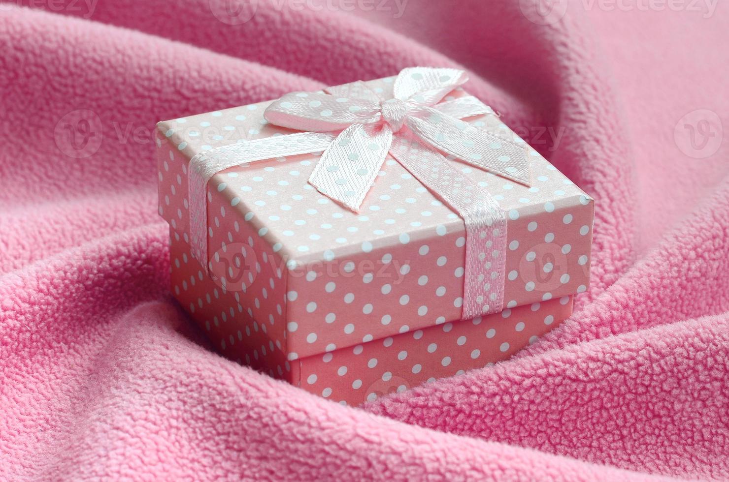 una pequeña caja de regalo de color rosa con un pequeño lazo se encuentra sobre una manta de tela suave y peluda de color rosa claro con muchos pliegues en relieve. embalaje para un regalo a su encantadora novia foto