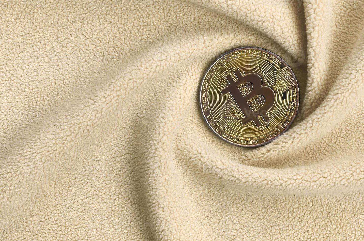 The golden bitcoin lies on a blanket made of soft and fluffy light orange fleece fabric with a large number of relief folds. The shape of the folds resembles a fan from a video card cooler photo