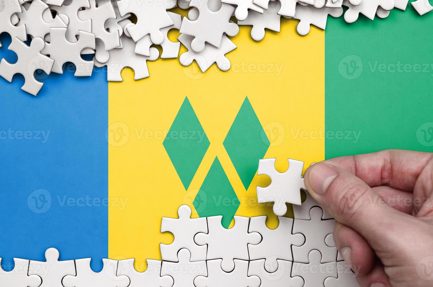 Saint Vincent and the Grenadines flag  is depicted on a table on which the human hand folds a puzzle of white color photo