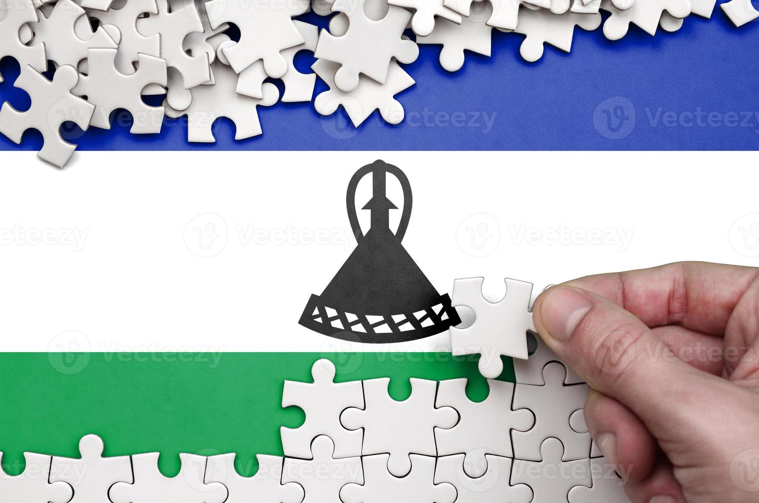 Lesotho flag  is depicted on a table on which the human hand folds a puzzle of white color photo