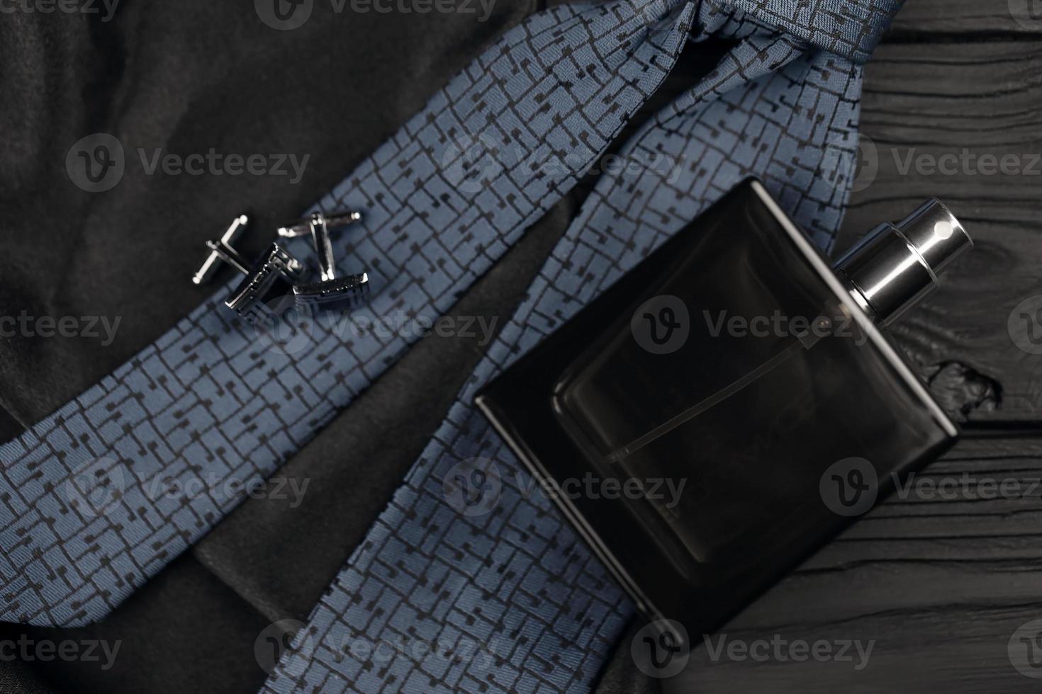 A bottle of mens cologne and cufflinks with blue tie lie on a black luxury fabric background on a wooden table. Mens accessories photo