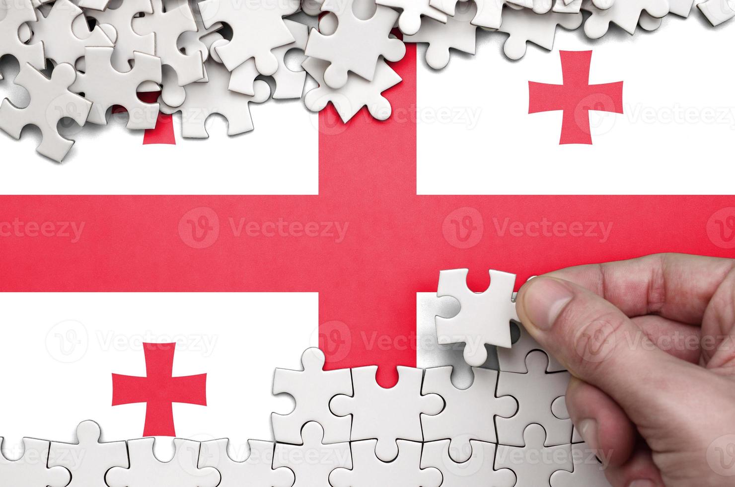 Georgia flag  is depicted on a table on which the human hand folds a puzzle of white color photo