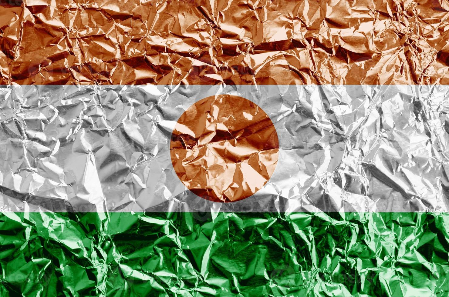 Niger flag depicted in paint colors on shiny crumpled aluminium foil closeup. Textured banner on rough background photo