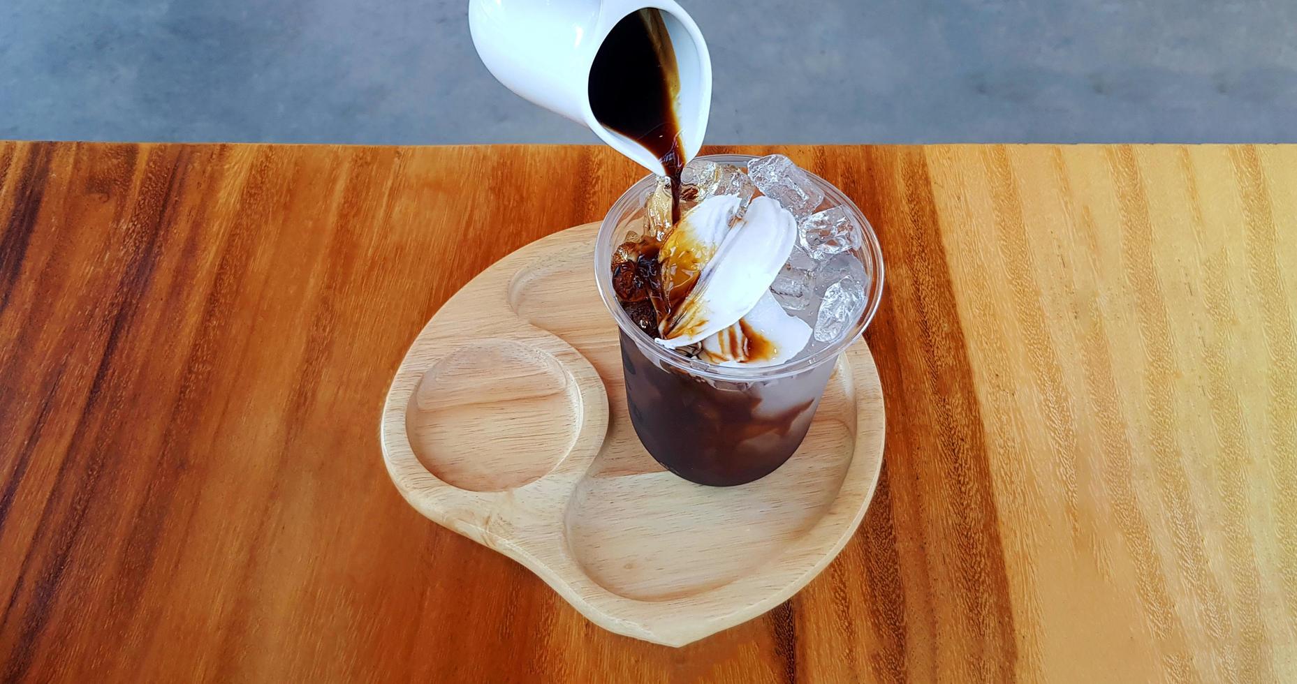 Pouring black or dark coffee in plastic glass of ice with fresh coconut on brown wooden table at cafe. Cold drinking and refreshment. Iced coconut coffee menu. photo
