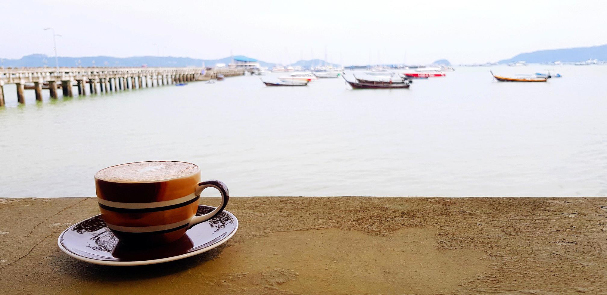 Brown cup of coffee on concrete table with seascape, ocean, bridge and boat background. Hot latte, coco or espresso with landscape view and copy space. Drinking and Relaxing concept. photo