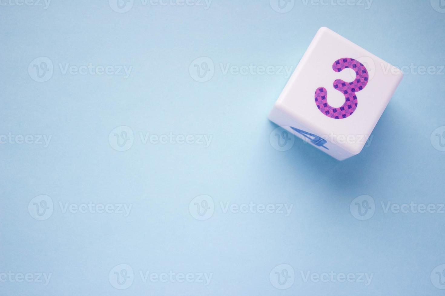 Close-up photo of a white plastic cube with a violet number 3 on a blue background in the upper right corner. With copy space