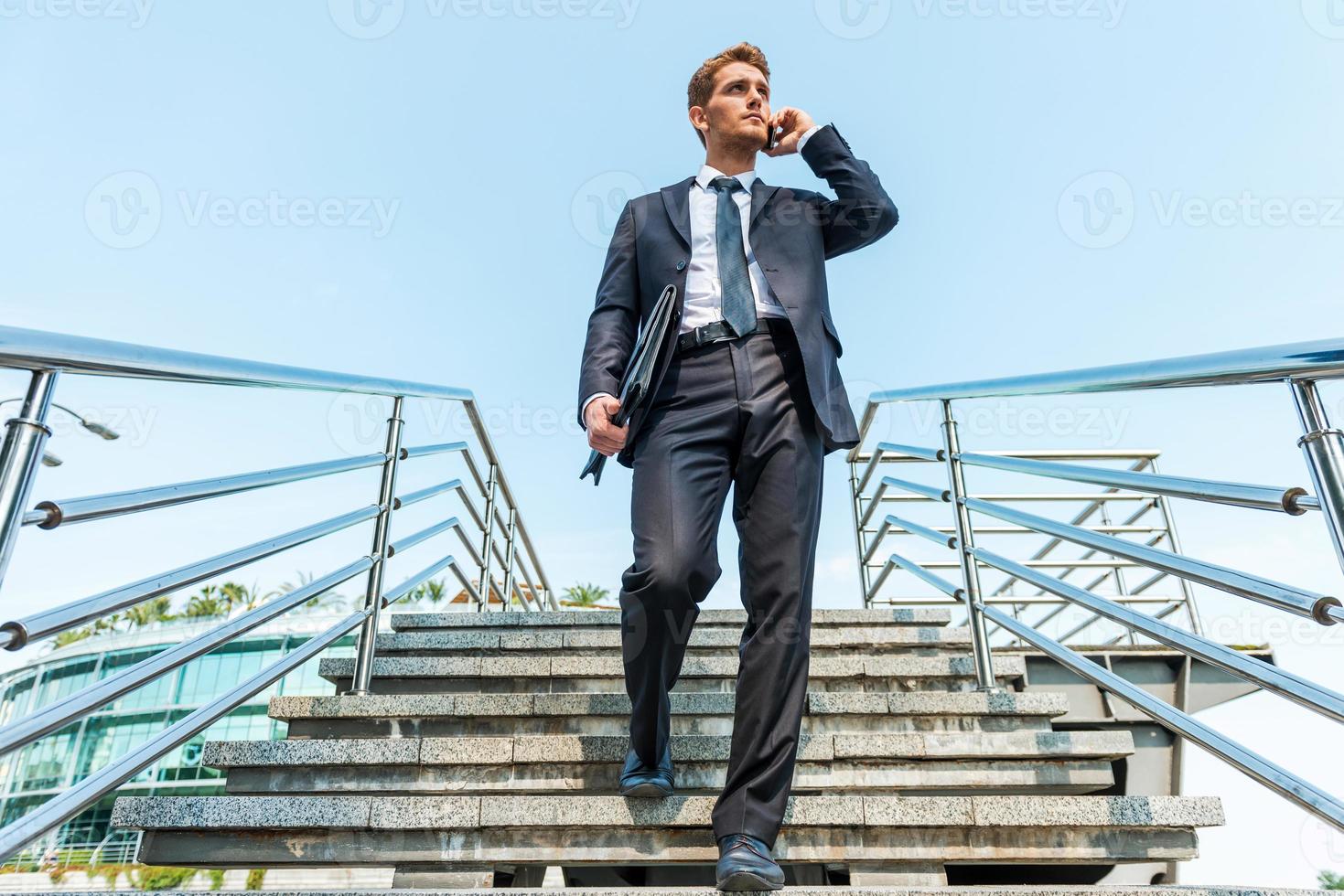 hombre de negocios confiado en el teléfono. vista de ángulo bajo de un joven confiado en ropa formal hablando por teléfono móvil mientras baja por la escalera foto