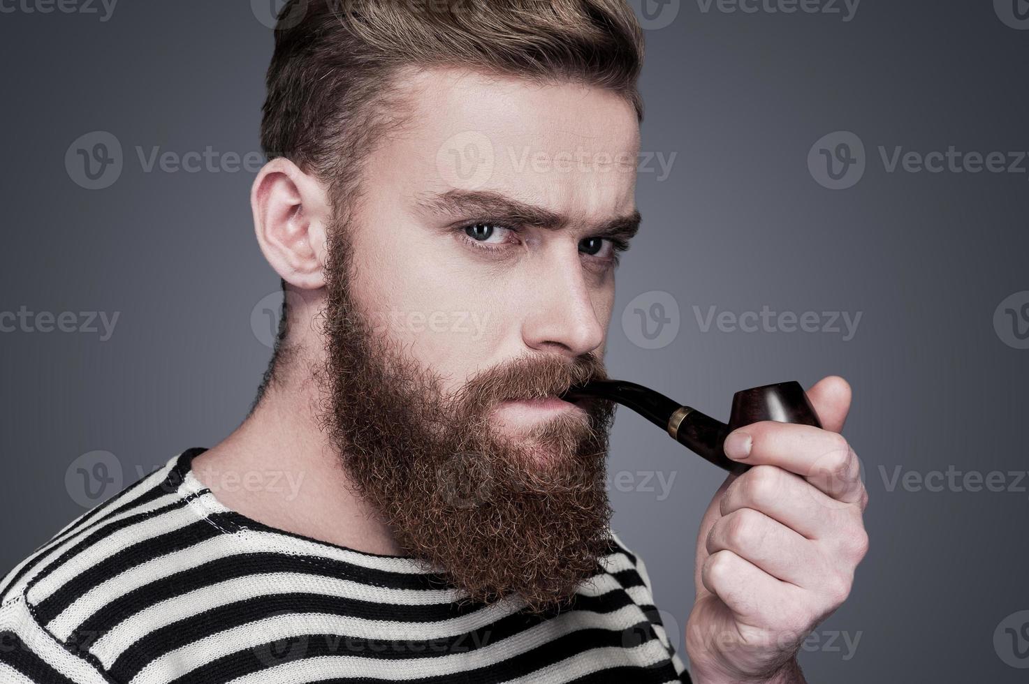 Sailor with pipe. Confident young bearded man in striped clothing smoking a pipe and looking at camera while standing against grey background photo