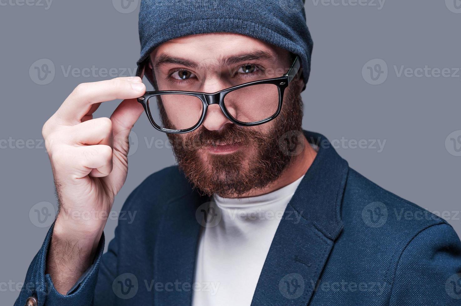 Hi there Handsome young bearded man in smart casual wear adjusting his eyeglasses and looking at camera while standing against grey background photo